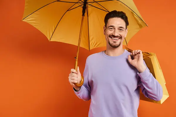 Homme gai avec parapluie jaune et sacs à provisions debout sur fond orange, consumérisme — Photo de stock