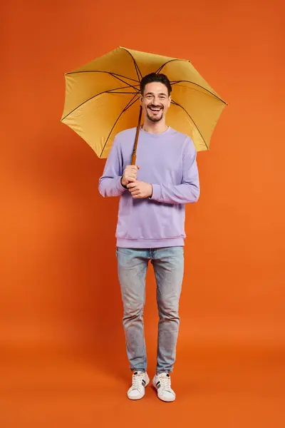 Cheerful man in purple sweatshirt standing with yellow umbrella on orange background, vibrant colors — Stock Photo