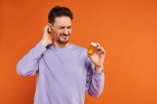 Bärtiger Mann in lila Sweatshirt mit Flasche mit Pillen auf orangefarbenem Hintergrund, Medikamente — Stockfoto