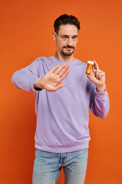 Homem barbudo em camisola roxa segurando garrafa com pílulas e mostrando parar no fundo laranja — Fotografia de Stock
