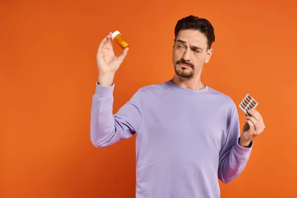 Bearded man in purple sweatshirt holding bottle with pills and blister pack on orange background — Stock Photo