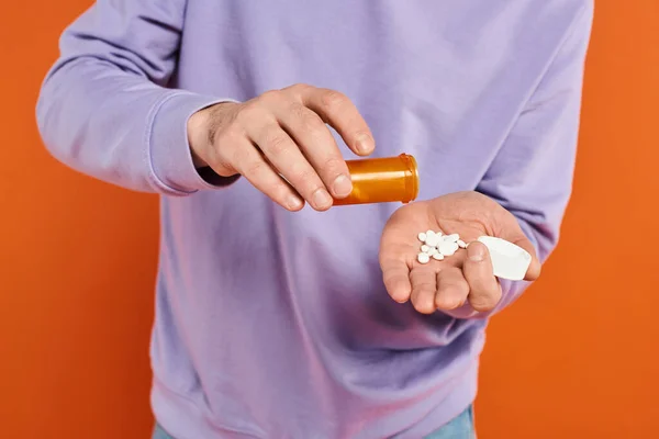 Homem barbudo em camisola roxa derramando pílulas na mão sobre fundo laranja, medicação — Fotografia de Stock