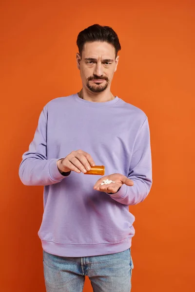 Homem barbudo em camisola roxa derramando medicação na mão sobre fundo laranja, pílulas — Fotografia de Stock