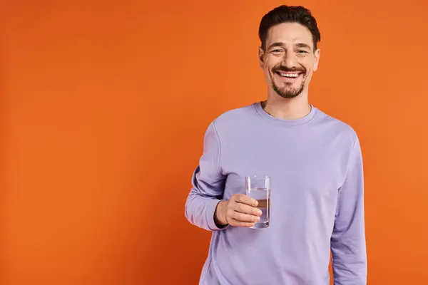 Happy bearded man in purple sweatshirt holding glass of water on orange background, hydration — Stock Photo