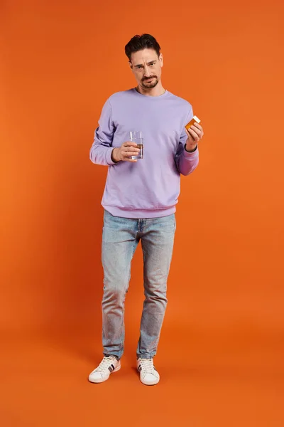 Бородатий чоловік у фіолетовому светрі тримає склянку води та пляшку з таблетками на помаранчевому фоні — стокове фото