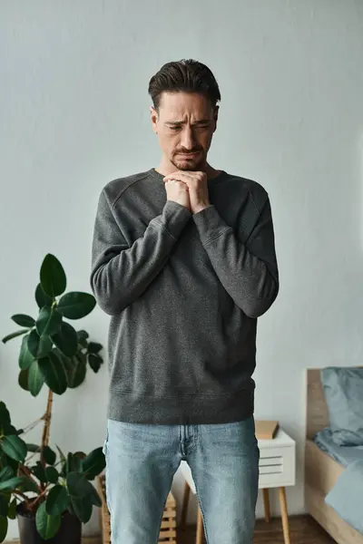 Homme déprimé et barbu en tenue de maison décontractée debout avec les mains près du visage sur fond gris, triste — Photo de stock