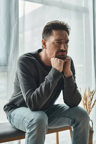 Hombre triste y barbudo en ropa casera casual sentado en el banco del dormitorio y mirando hacia otro lado, depresión - foto de stock