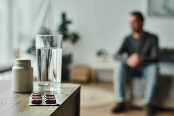 Medication in bottle and blister pack near glass of water on table and blurred man on background — Stock Photo