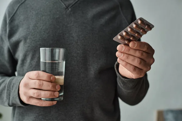 Cropped photo of man in casual grey jumper holding glass of water and blister pack with medication — Stock Photo