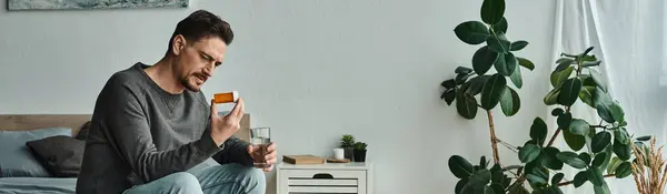 Banner of bearded man holding glass of water while looking at medication and sitting on bed — Stock Photo