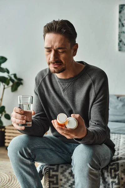 Bearded man holding glass of water while looking at medication in bottles and sitting on bed — Stock Photo