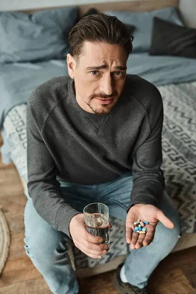 Bärtiger Mann runzelt die Stirn, während er ein Glas Wasser und Medikamente in den Händen hält und in die Kamera blickt — Stockfoto