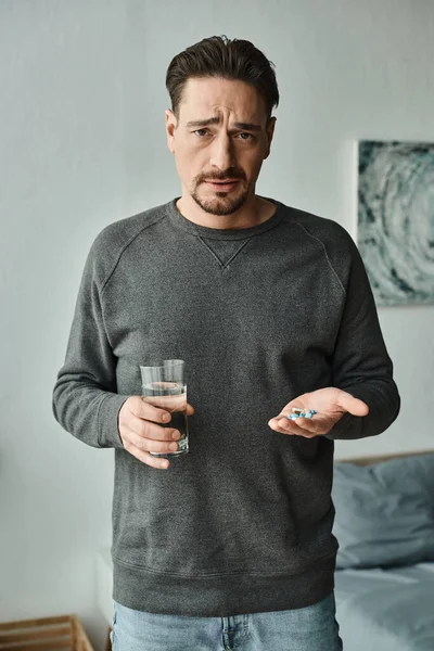 Bärtiger Mann, der in die Kamera schaut, während er ein Glas Wasser und Medikamente in den Händen hält — Stockfoto