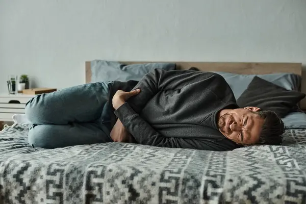 Uomo barbuto sdraiato sul letto e accigliato dal dolore mentre soffre di mal di stomaco, smorfia e dolore — Foto stock