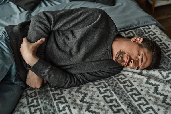 Hombre barbudo haciendo muecas mientras yacía en la cama y sufría de dolor de estómago, dolor abdominal - foto de stock
