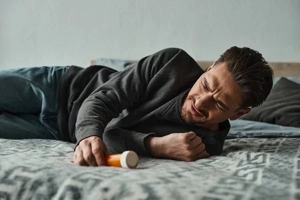 Homme barbu souffrant de douleur et couché sur le lit près de la bouteille avec des médicaments, des crampes et de l'inconfort — Photo de stock