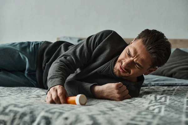 Bearded man suffering from pain and lying on bed near bottle with medication, cramp and discomfort — Stock Photo
