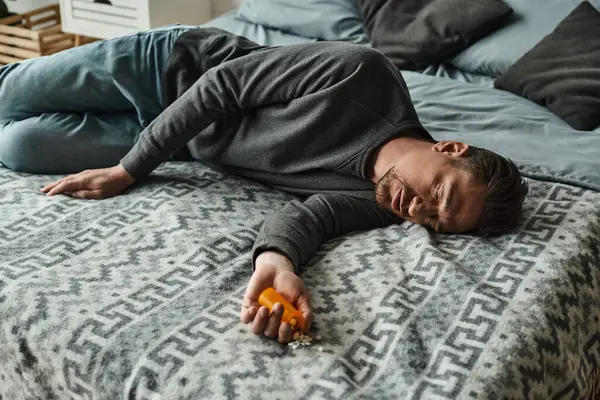 Homme barbu souffrant de douleur et regardant bouteille avec des pilules tout en étant couché sur le lit avec couverture — Photo de stock