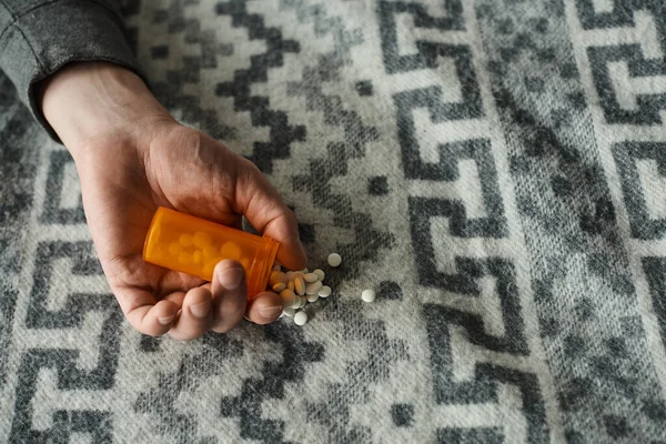 Vue recadrée de l'homme tenant bouteille avec des pilules déversées sur la couverture grise avec ornement, médicaments — Photo de stock