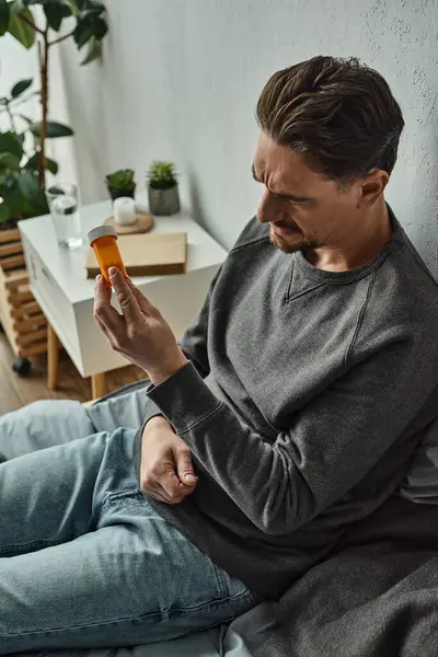 Homem pensativo examinando garrafa com medicação enquanto sentado na cama no quarto, plano de tratamento — Fotografia de Stock