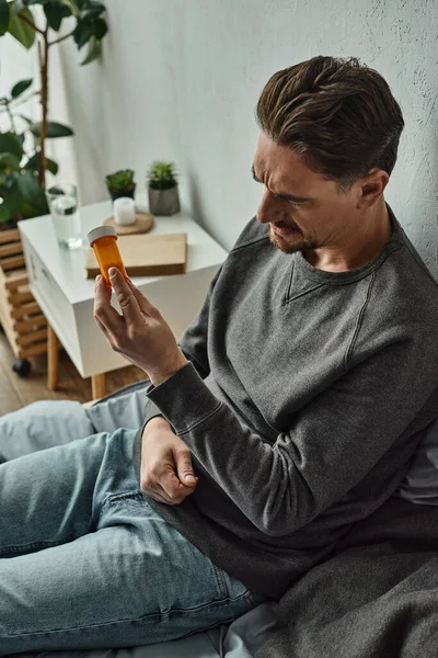 Pensive man examining bottle with medication while sitting on bed in bedroom, treatment plan — Stock Photo