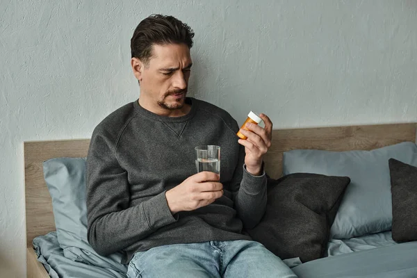 Uomo barbuto in maglione con bicchiere d'acqua e bottiglia con pillole in camera da letto, trattamento — Foto stock