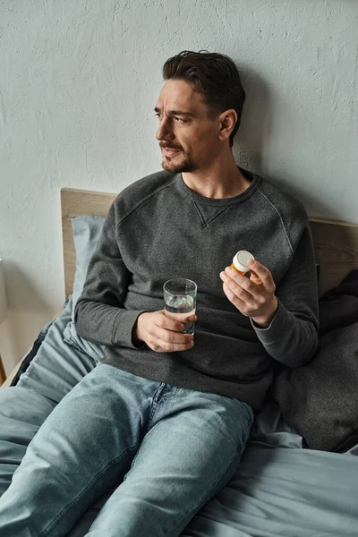 Uomo premuroso in maglione che tiene bicchiere d'acqua e bottiglia con pillole in camera da letto, trattamento — Foto stock