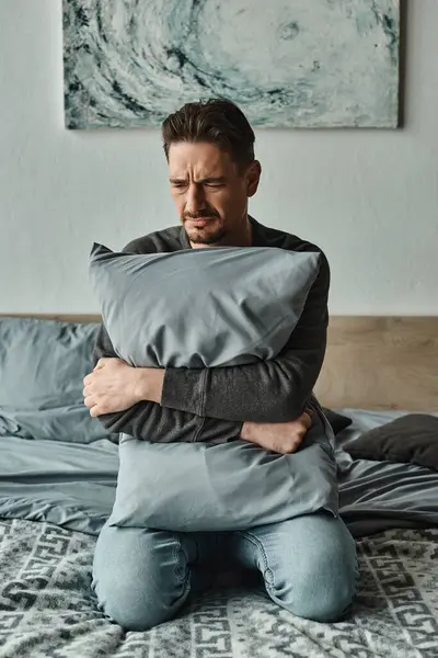 Bearded man feeling discomfort and holding pillow while sitting on bed at home, troubled patient — Stock Photo