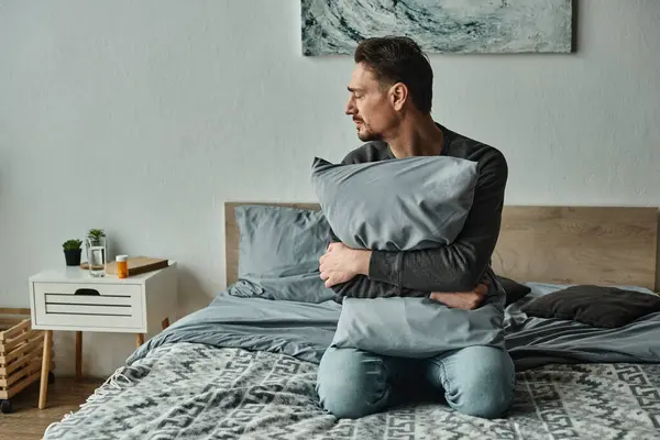 Бородатий чоловік відчуває дискомфорт і тримає подушку, сидячи на ліжку вдома, сучасний інтер'єр — стокове фото