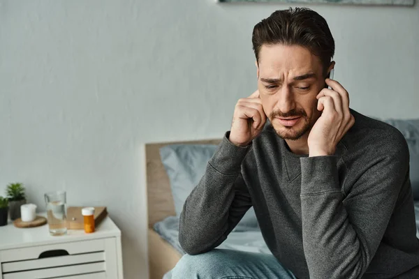 Homem barbudo com dor de cabeça falando no smartphone no quarto moderno, consulta on-line — Fotografia de Stock