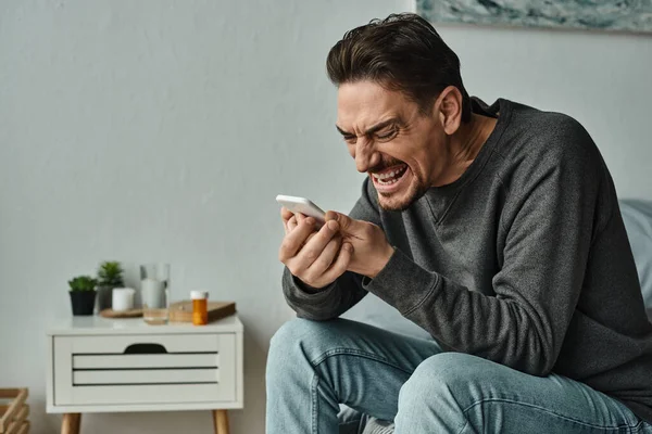 Uomo barbuto in maglione grigio urlando al suo smartphone mentre seduto sul letto a casa, emotivo — Foto stock
