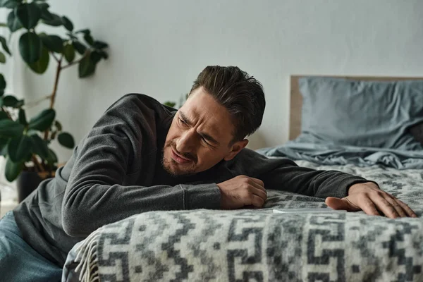 Homem barbudo sofrendo de dor e apoiando-se em cobertor cinza na cama no quarto moderno, estresse — Fotografia de Stock
