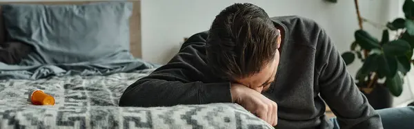 Uomo barbuto che soffre di dolore e appoggiato sulla coperta grigia sul letto in camera da letto moderna, banner dolore — Foto stock