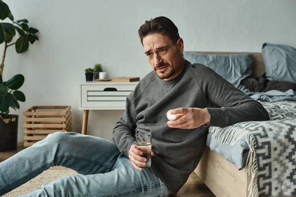 Hombre barbudo preocupado sentado en el suelo y sosteniendo un vaso de agua y pastillas en el dormitorio, medicamentos - foto de stock