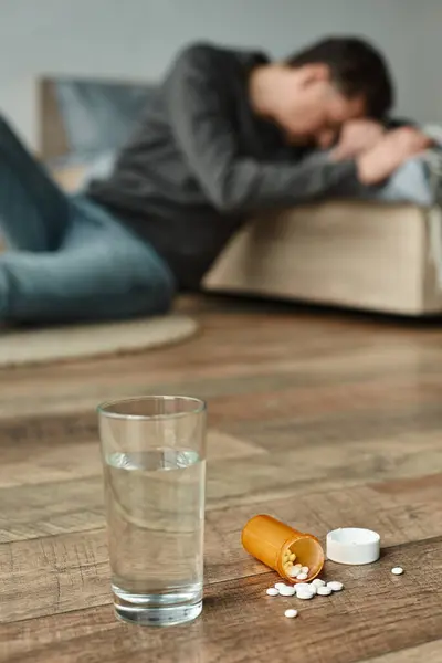 Vaso de agua y botella con pastillas en el suelo cerca del hombre borroso que sufre de dolor en el fondo - foto de stock