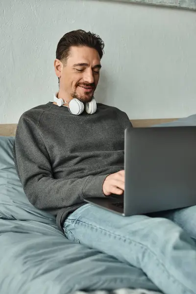 Щасливий чоловік у бездротових навушниках, використовуючи ноутбук, сидячи на ліжку, позаштатний та віддалений робочий — стокове фото