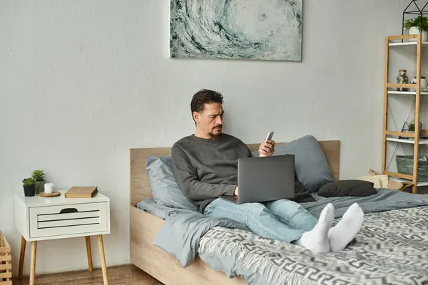 Homem barbudo usando laptop e segurando telefone celular enquanto trabalhava remotamente de casa, freelance — Fotografia de Stock