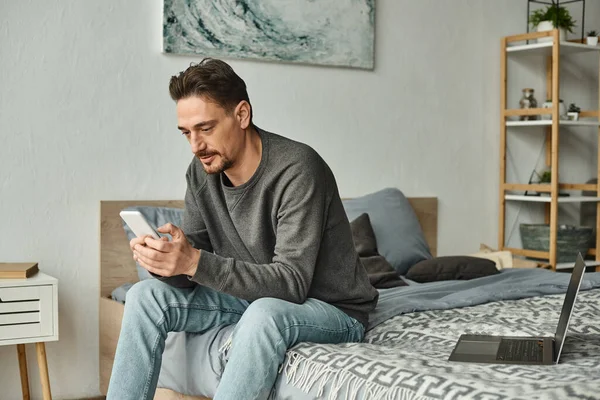 Barbu homme textos sur téléphone mobile tout en travaillant à distance de la maison près d'un ordinateur portable sur le lit, freelance — Photo de stock