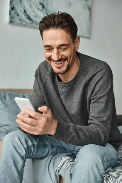 Hombre barbudo alegre en suéter gris mensajes de texto en el teléfono inteligente en el dormitorio moderno, las redes sociales - foto de stock