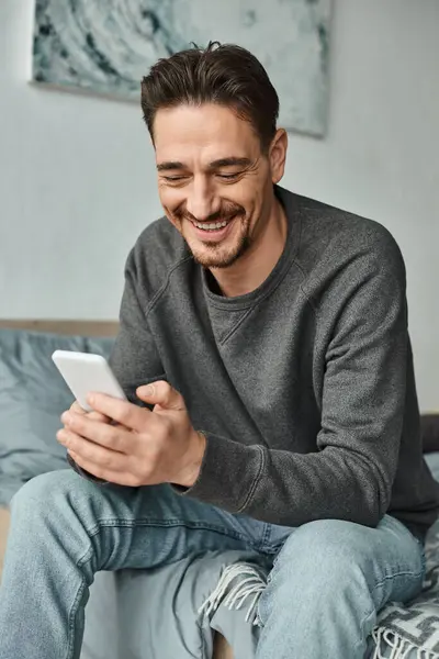 Allegro uomo barbuto in maglione grigio sms su smartphone in camera da letto moderna, social media — Foto stock