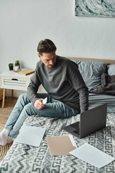 Focused bearded man holding smartphone and using laptop near documents on bed, remote work — Stock Photo