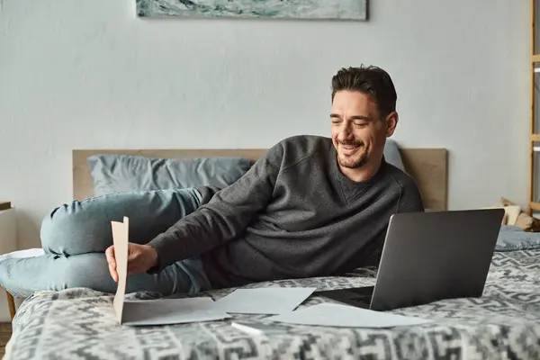 Fröhlicher und bärtiger Mann mit Laptop, während er von zu Hause aus im Schlafzimmer arbeitet, Dokumente — Stockfoto
