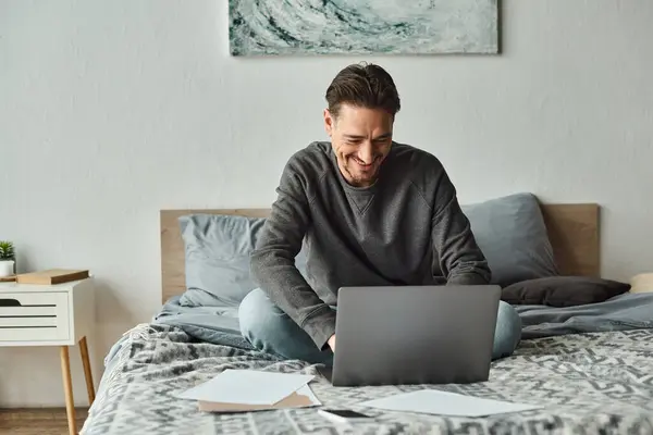 Uomo gioioso e barbuto utilizzando il computer portatile mentre si lavora in remoto da casa in camera da letto, documenti — Foto stock