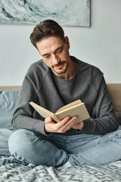 Bärtiger Mann im grauen lässigen Pullover liest Buch, während er am Wochenende im Schlafzimmer entspannen, Freizeit — Stockfoto