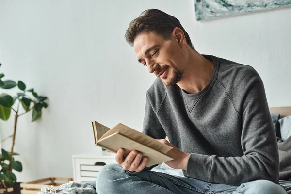 Zufriedener Mann im grauen lässigen Pullover, der Buch liest, während er am Wochenende im Schlafzimmer entspannen kann, Freizeit — Stockfoto