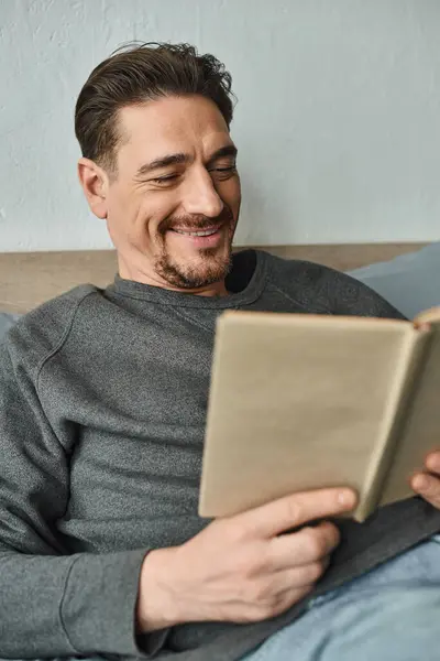 Homme gai en gris casual pull livre de lecture tout en se relaxant le week-end dans la chambre, loisirs — Photo de stock