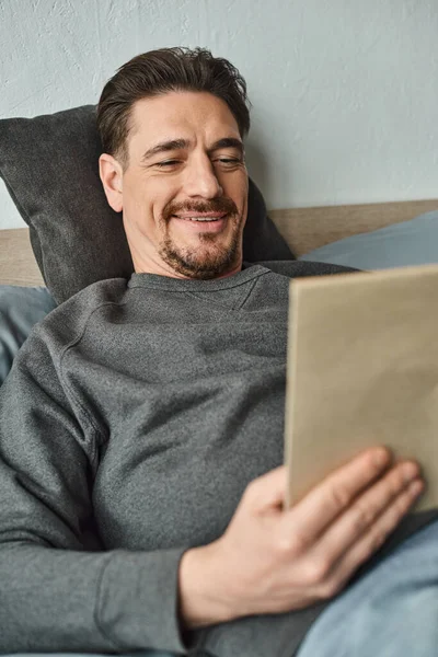 Cheerful man in casual jumper reading book while relaxing on weekend in bedroom, leisure concept — Stock Photo