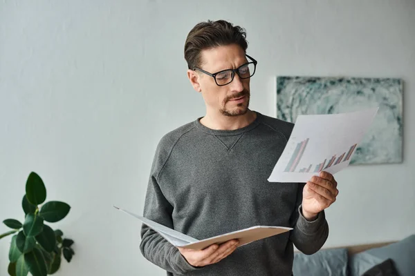 Focused and bearded man in eyeglasses and grey sweater analyzing graphs while working from home — Stock Photo
