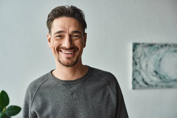 Portrait of cheerful bearded man in grey jumper looking at camera on bedroom, satisfaction — Stock Photo