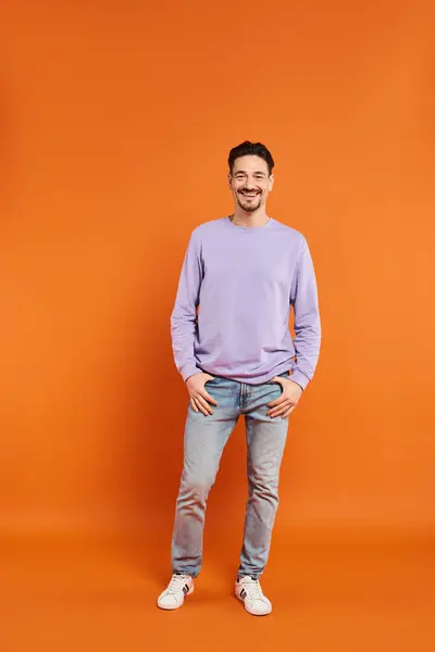 Longitud completa de hombre alegre y barbudo en suéter púrpura mirando a la cámara sobre fondo naranja - foto de stock